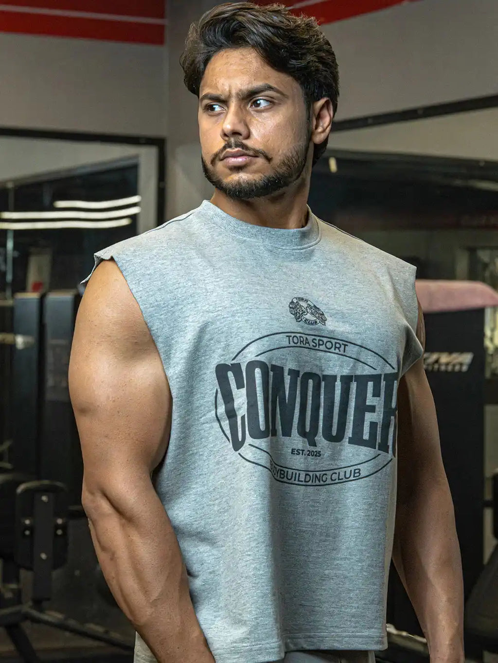 Muscular man in sleeveless grey gym shirt with 'Conquer' print posing in fitness center