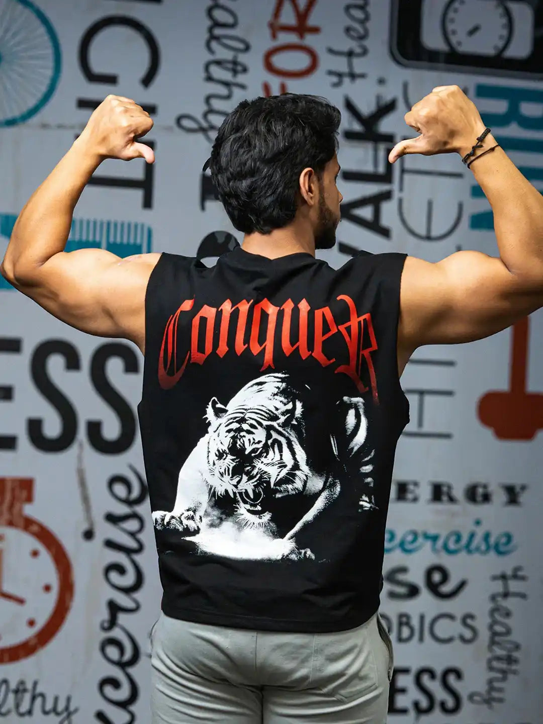 Man flexing in gym wearing black sleeveless shirt with roaring tiger and 'Conquer' text on back