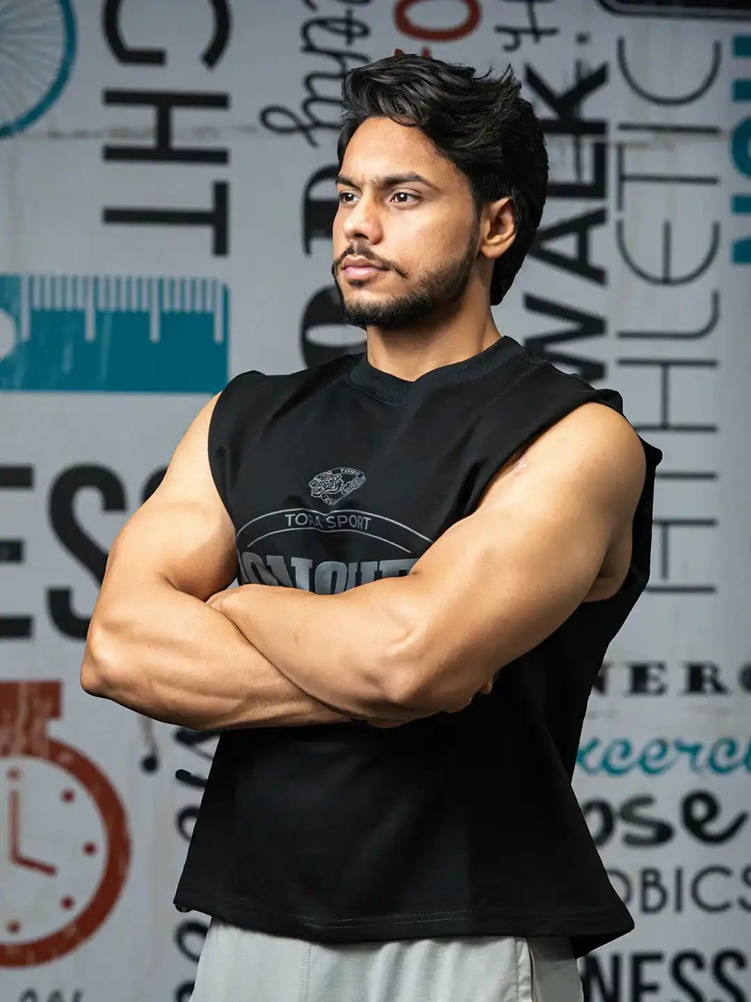 Athletic man in black sleeveless gym shirt with arms crossed, fitness-themed wall background.