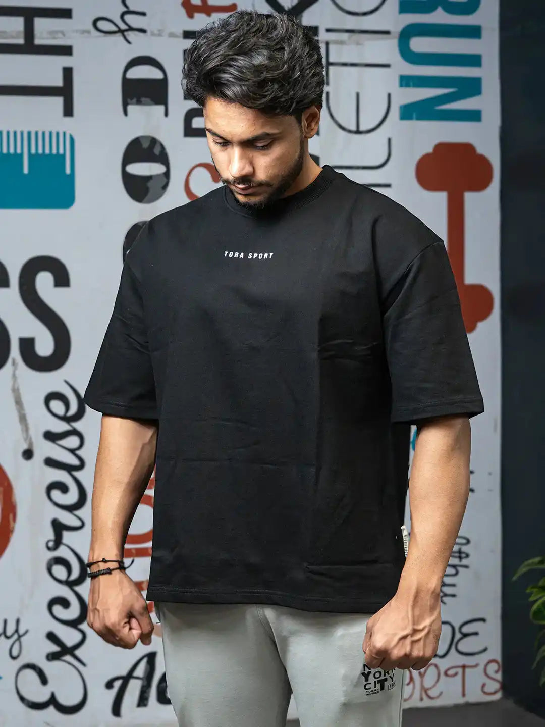 Man wearing black oversized T-shirt with Tora Sport logo, gym wear style indoors.
