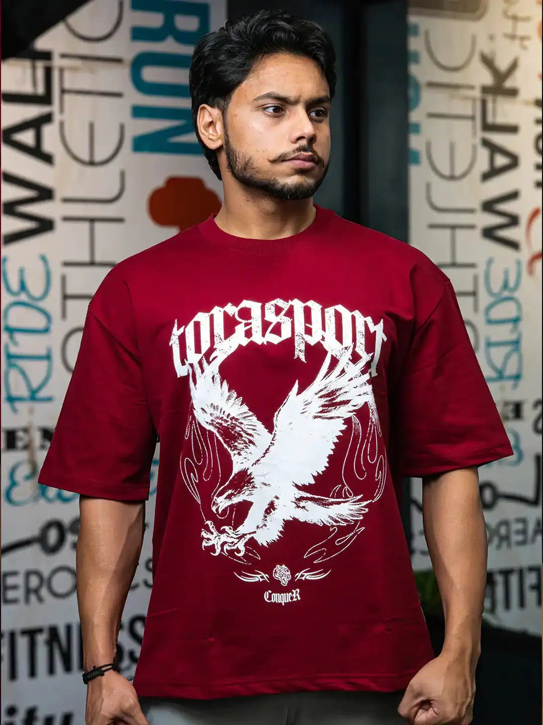Man wearing maroon gym t-shirt with bold eagle graphic and 'Conquer' text, urban backdrop.