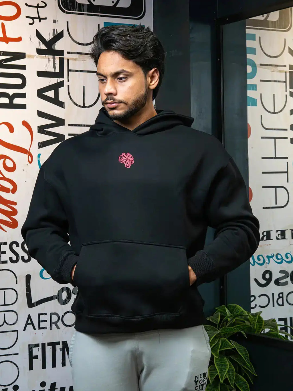 Man in black gym hoodie with front pocket and red logo, standing indoors by motivational wall.