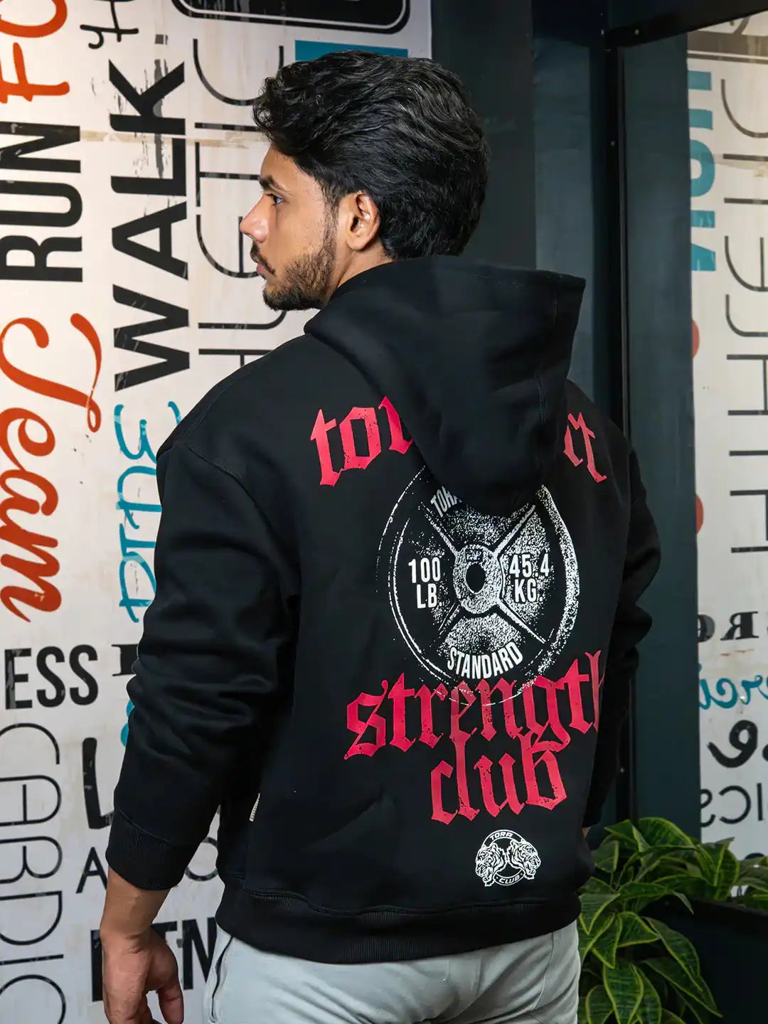 Man wearing black hoodie with weight plate and red 'strength club' graphic, gym wall art background