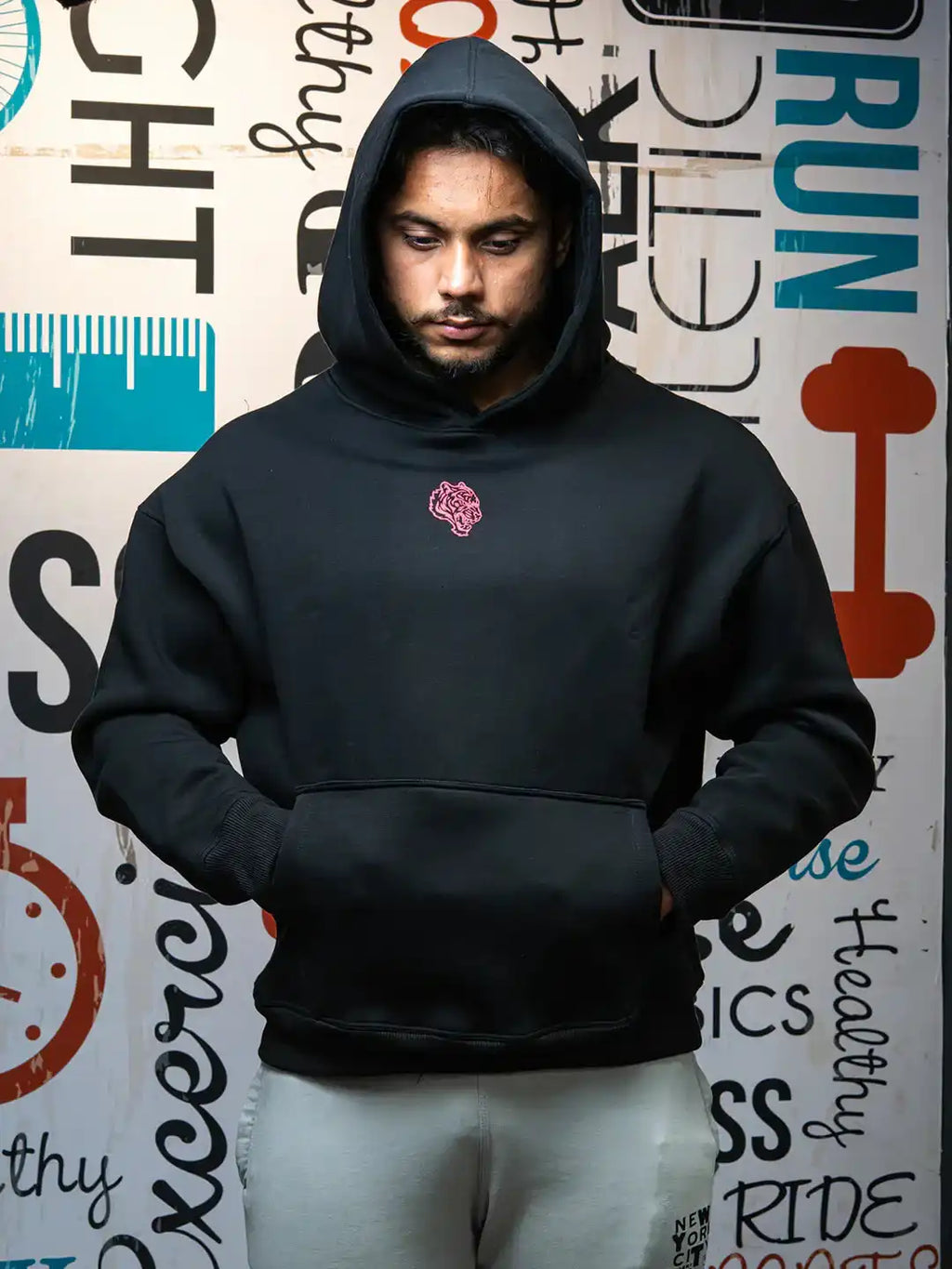 Man wearing black gym hoodie with tiger emblem, hands in pocket, standing indoors