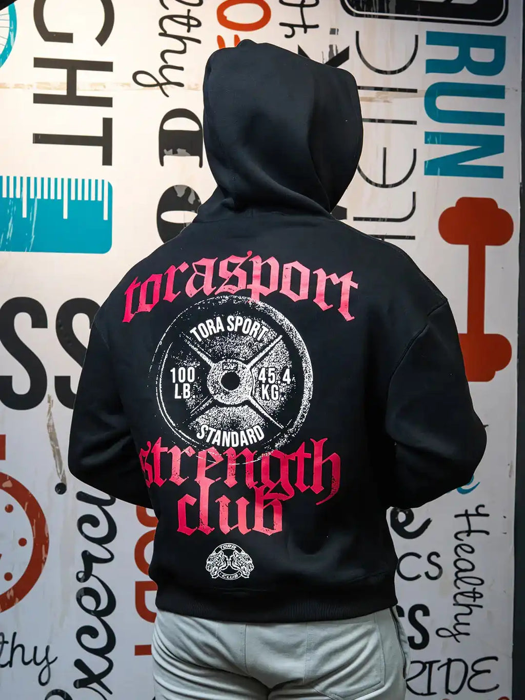 Person wearing black hoodie with bold gym-themed graphic and text 'strength club' on back, standing by gym wall.